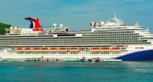 Large white cruise ship with multiple decks and red funnel docked at a port, with clear blue sky and lush green trees in the background. People are visible walking along the dock.