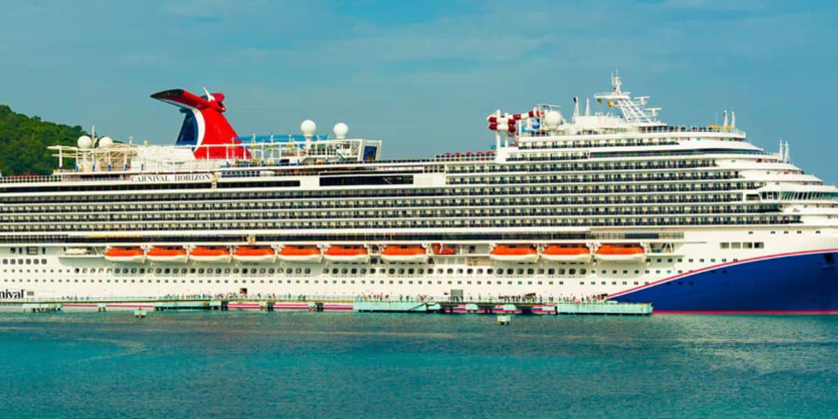Large white cruise ship with multiple decks and red funnel docked at a port, with clear blue sky and lush green trees in the background. People are visible walking along the dock.