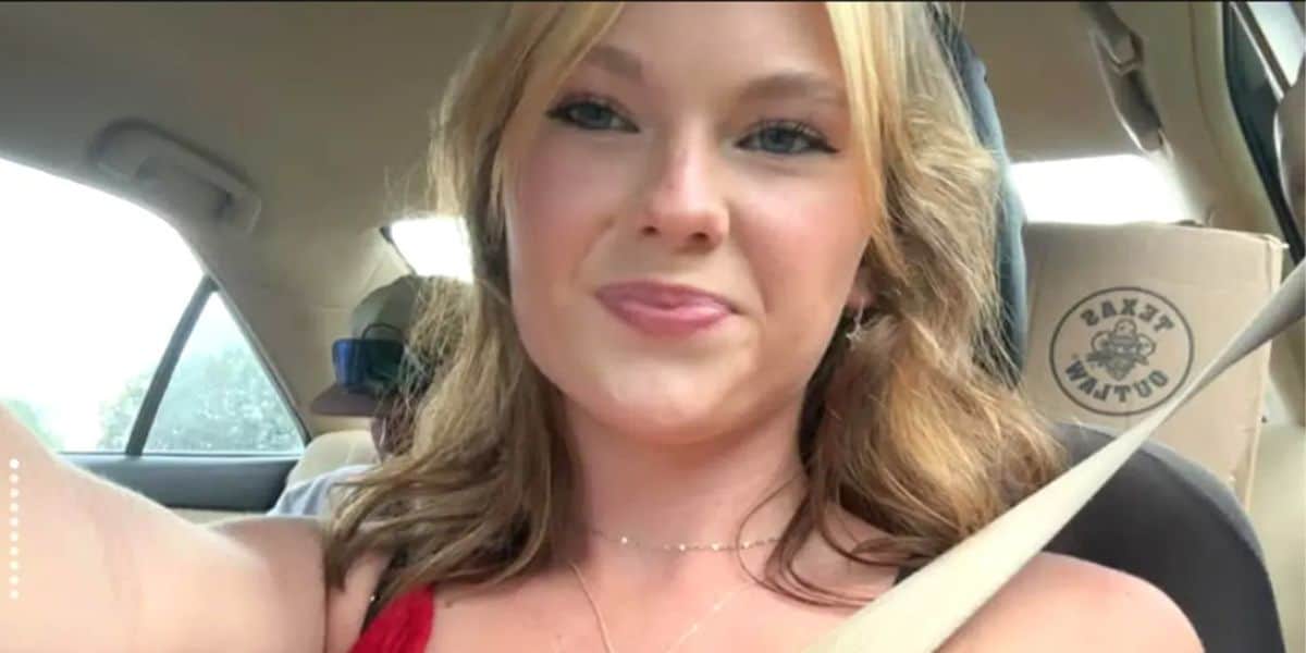 A young woman with blonde hair smiles slightly while taking a selfie in a car. She is wearing a red top and a necklace, and sunlight brightens her face. The car seat headrest has a logo that reads “SAXY MAFIA.”.