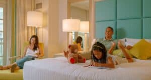A family unwinds in a bright Disney hotel room; a girl sketches on the bed while parents read after a fun day at the parks.