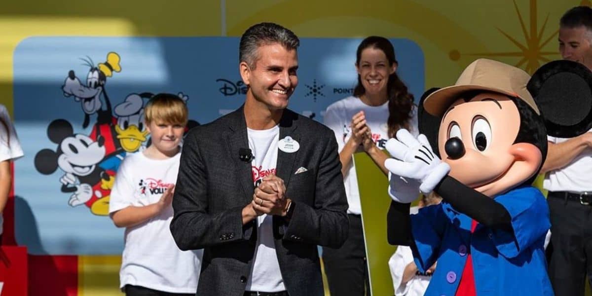 Mickey Mouse and an executive share applause on stage with Disney fans, set against a vibrant Disney World backdrop.