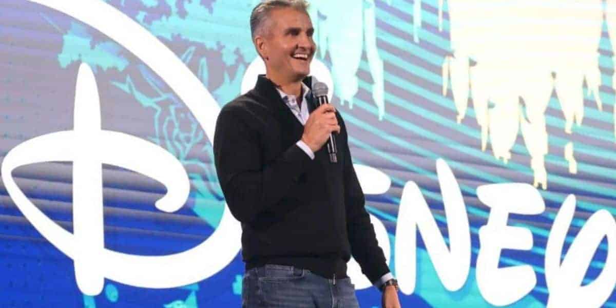 A Disney presenter grins onstage, microphone in hand, with the iconic Disney logo glowing large behind him at a live event.