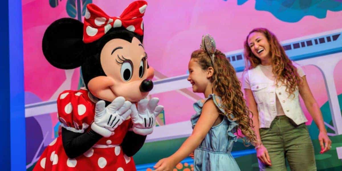 A young guest smiles meeting Minnie Mouse at Disney World, with families enjoying a vibrant park atmosphere in the background.