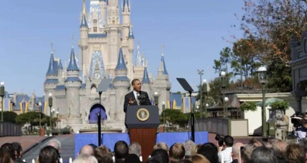 A Disney Parks host stands at a podium before Cinderella Castle as fans and media capture the high-energy celebration outdoors.