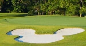 A Mickey Mouse-shaped sand bunker by the green at a Disney World golf course, surrounded by lush trees.