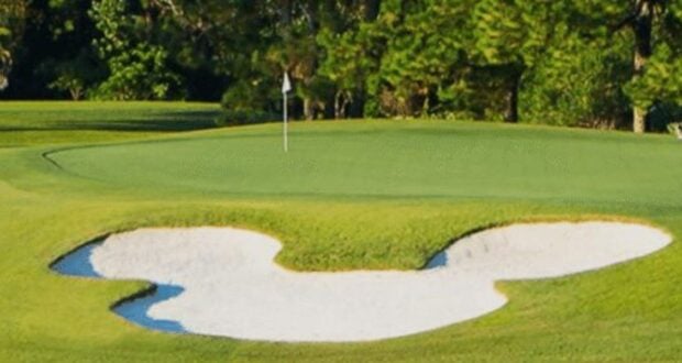 A Mickey Mouse-shaped sand bunker by the green at a Disney World golf course, surrounded by lush trees.