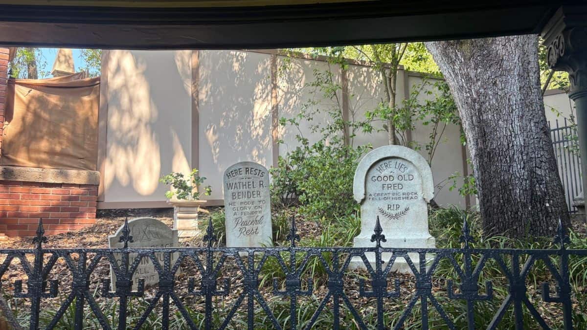 Haunted Mansion tombstones behind a black fence, set among trees and brick walls, evoke Tomorrowland’s mysterious vibe.