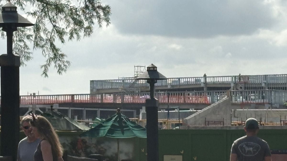 Excitement builds at Disney’s Animal Kingdom as green fencing reveals a future Encanto attraction; guests stroll with umbrellas.