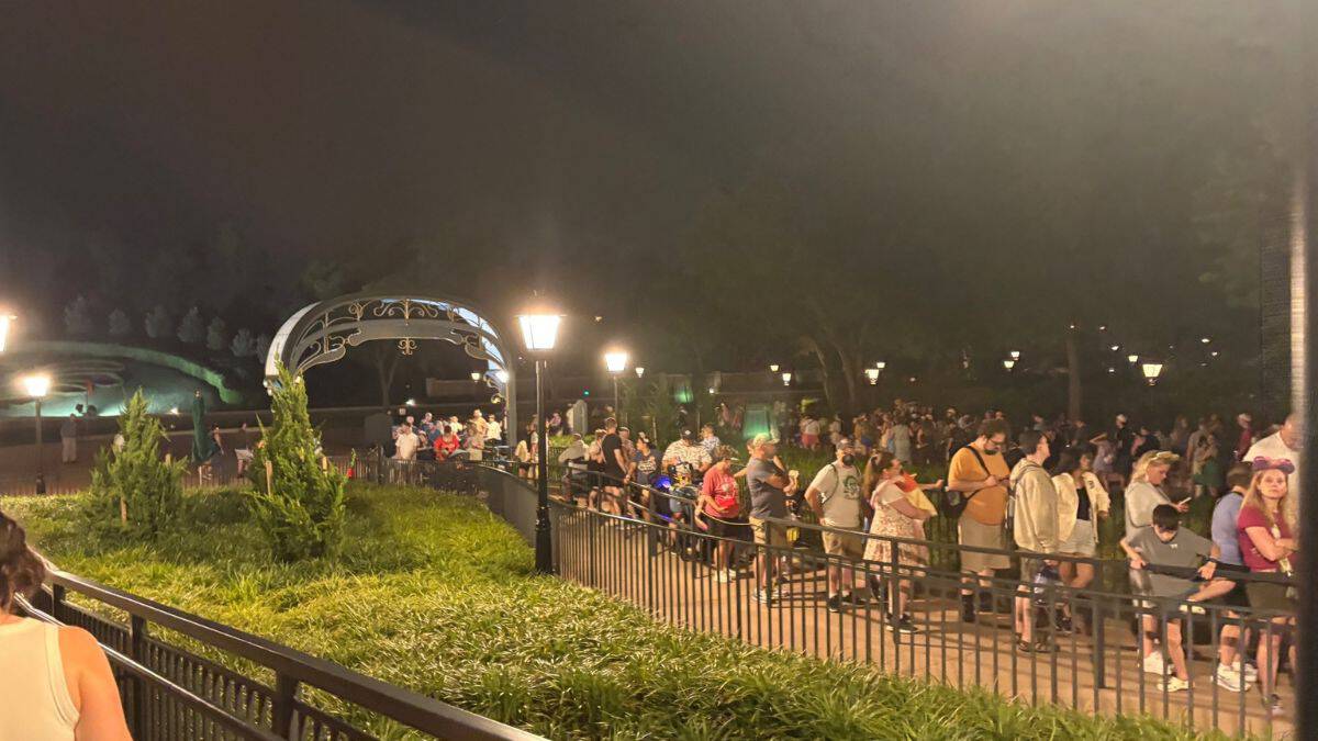Guests queue in a serpentine line at night near Disney World's glowing arch and landscaped lawnsโclassic Skyliner crowds at close.