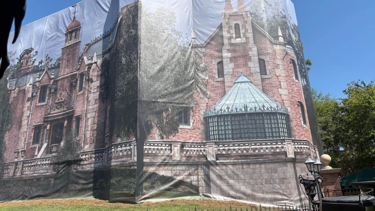 A building is wrapped in a tarp showing a grand brick mansion with towers, giving it Tomorrowland vibes that draw park guests’ attention.