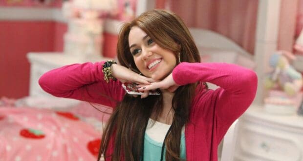 A teen girl in a pink cardigan strikes a playful Disney Channel pose, hands under chin, in a vibrant bedroom setting.