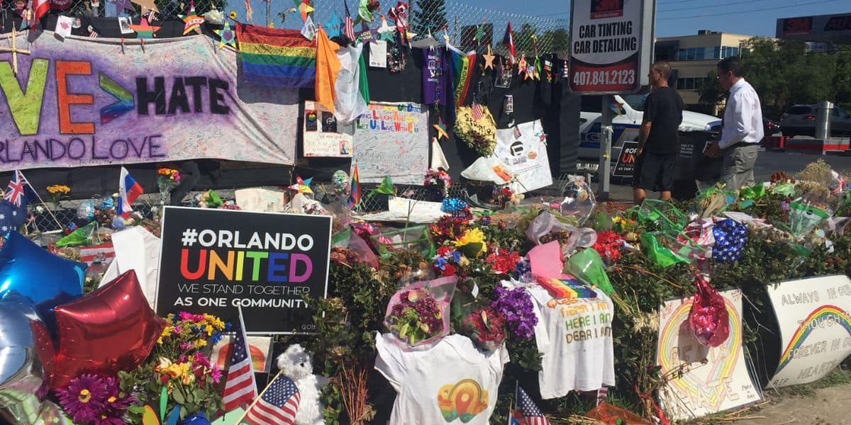 A tribute of flowers, signs, and rainbow flags remembers Pulse victims and notes the shooter's connection to Disney World.