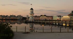 A glowing boardwalk lighthouse with illuminated entertainment venues reflecting on tranquil water at dusk.