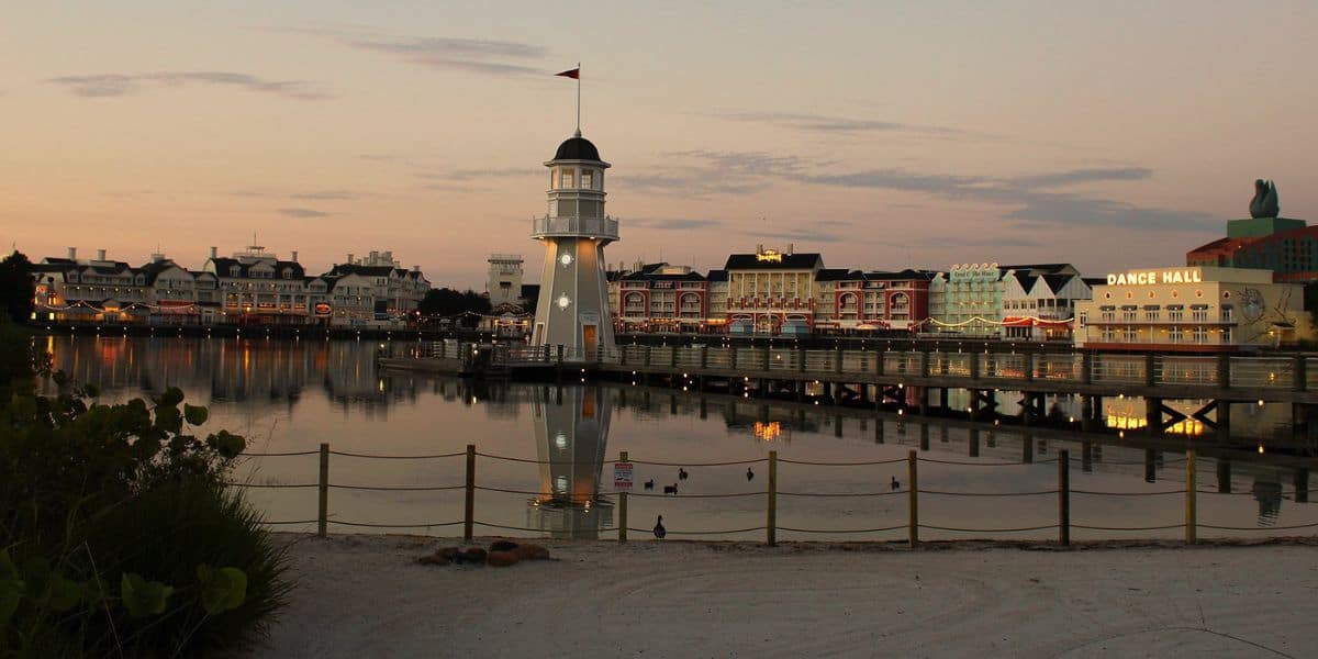 A glowing boardwalk lighthouse with illuminated entertainment venues reflecting on tranquil water at dusk.