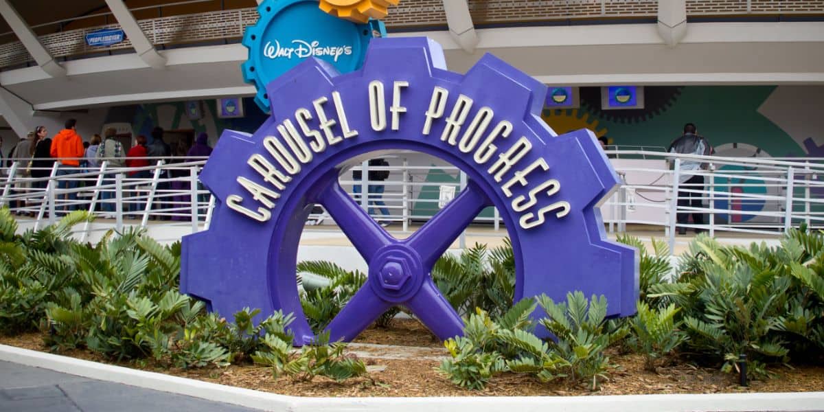The iconic purple gear-shaped Carousel of Progress sign welcomes guests amid lush greenery at the entrance to Disney World.