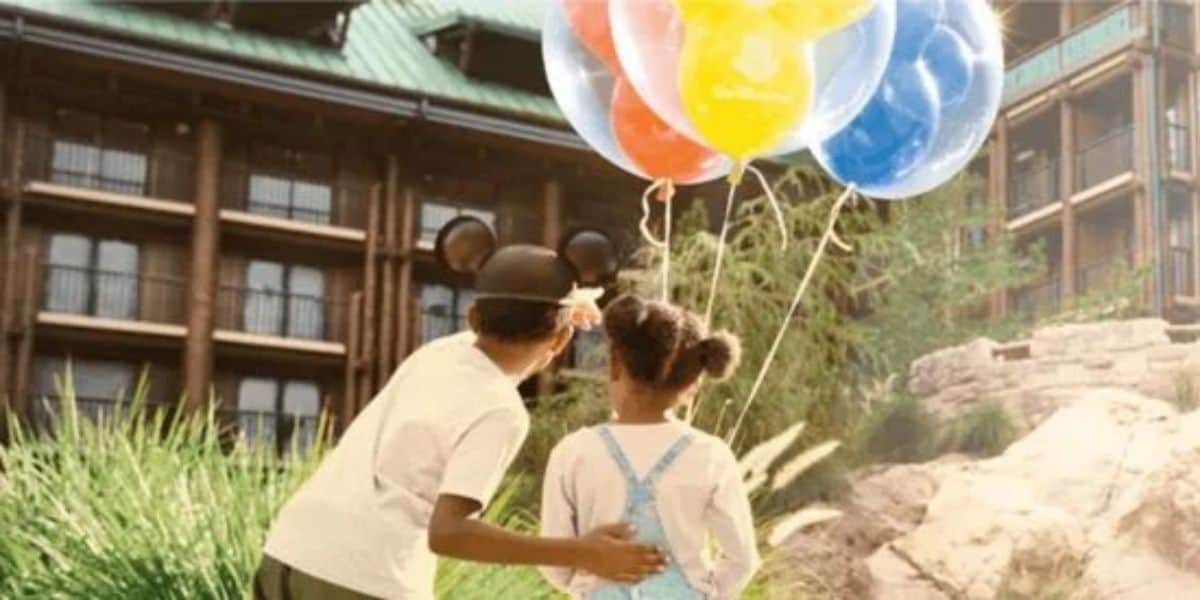 Two children stand together outside, gazing up at a bunch of colorful balloons. The child on the left sports mouse ears, reminiscent of a Disney World guest. Sunlight illuminates the scene, highlighting nearby greenery against the backdrop of balcony-adorned buildings.