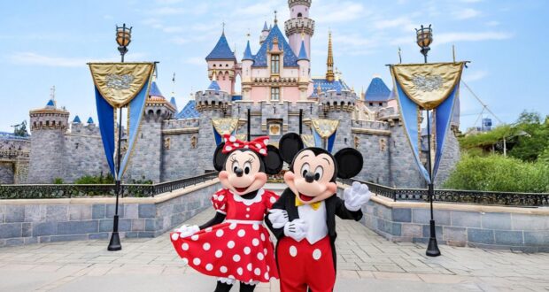 Mickey and Minnie Mouse greet guests in front of Disneyland’s iconic pink castle, adorned with sparkling blue rooftops and shimmering gold banners.