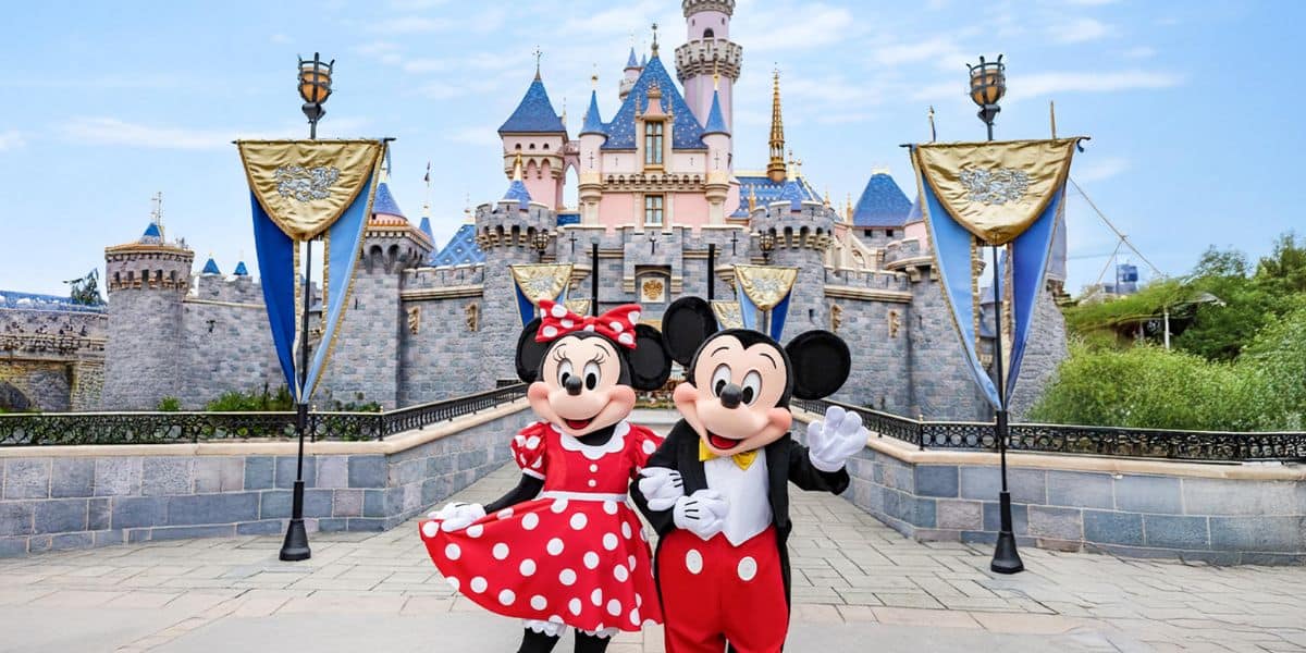 Mickey and Minnie Mouse greet guests in front of Disneyland’s iconic pink castle, adorned with sparkling blue rooftops and shimmering gold banners.