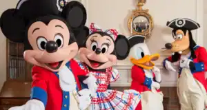 Mickey, Minnie, Donald, and Goofy greet guests in patriotic colonial outfits at a lively indoor Disney Parks celebration.