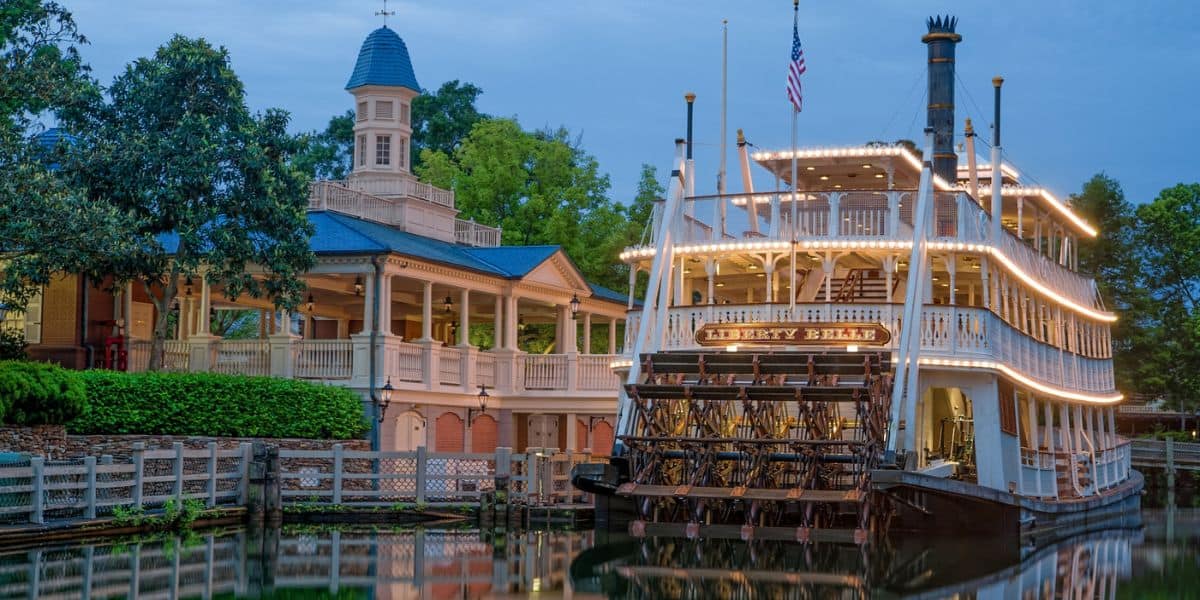 The illuminated riverboat docks near a rustic lodge and trees at dusk—echoing Rivers of America's farewell at Magic Kingdom.