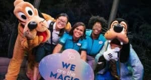 A group photo of three Disney cast members standing with Pluto and Goofy. The smiling cast members are holding a circular sign that reads, "We are the magic." They are outdoors, with trees and lights in the crisp background, truly showing what it means to be part of the Disney enchantment.