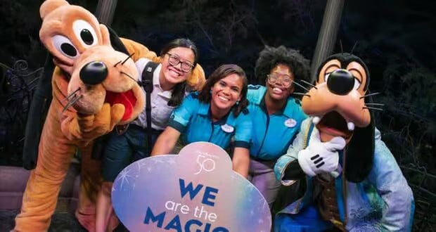 A group photo of three Disney cast members standing with Pluto and Goofy. The smiling cast members are holding a circular sign that reads, "We are the magic." They are outdoors, with trees and lights in the crisp background, truly showing what it means to be part of the Disney enchantment.