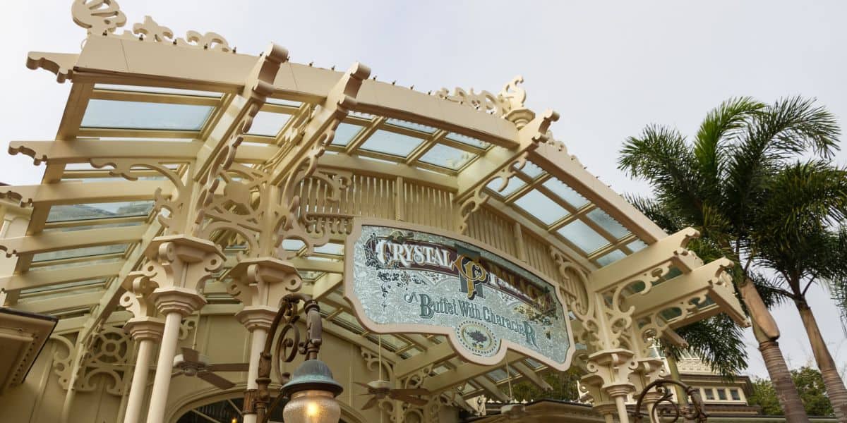 The iconic Crystal Palace entrance sign, adorned with ornate glass and cream details, welcomes guests amid lush palm trees in Magic Kingdom.