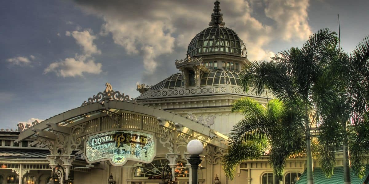 Grand entrance of an opulent glass pavilion at a theme park, framed by tropical palm trees and a vibrant marquee sign, set against dramatic, storybook-like clouds.