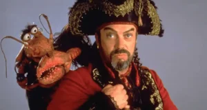 Captain Long John Silver, decked out in his signature pirate attire and channeling the iconic charm of Tim Curry, strikes a pose with the mischievous red Muppet, Clueless Morgan, perched on his shoulder—bringing classic Disney adventure to life against a simple backdrop.
