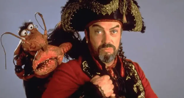 Captain Long John Silver, decked out in his signature pirate attire and channeling the iconic charm of Tim Curry, strikes a pose with the mischievous red Muppet, Clueless Morgan, perched on his shoulder—bringing classic Disney adventure to life against a simple backdrop.