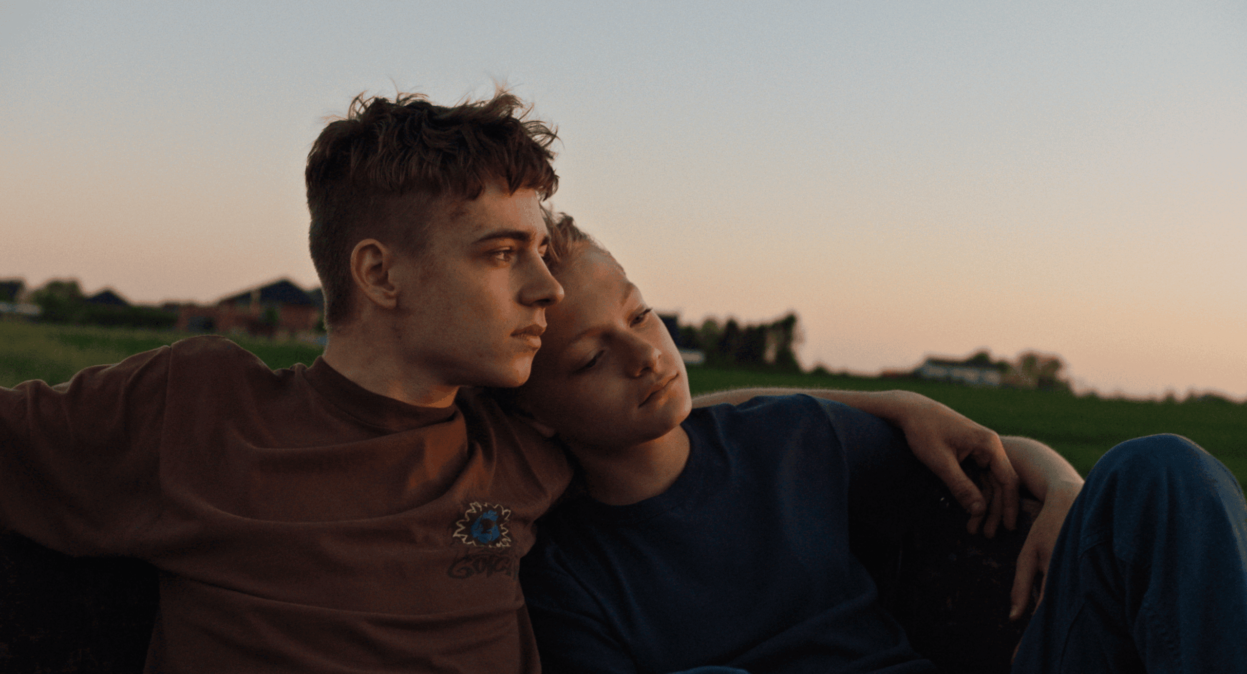 Dreamy outdoor scene with two teenage boys sitting closely, relaxed and contemplative, during sunset in a green field, emphasizing youth, friendship, and cinematic post-production quality for DIVO Post Production SEO.