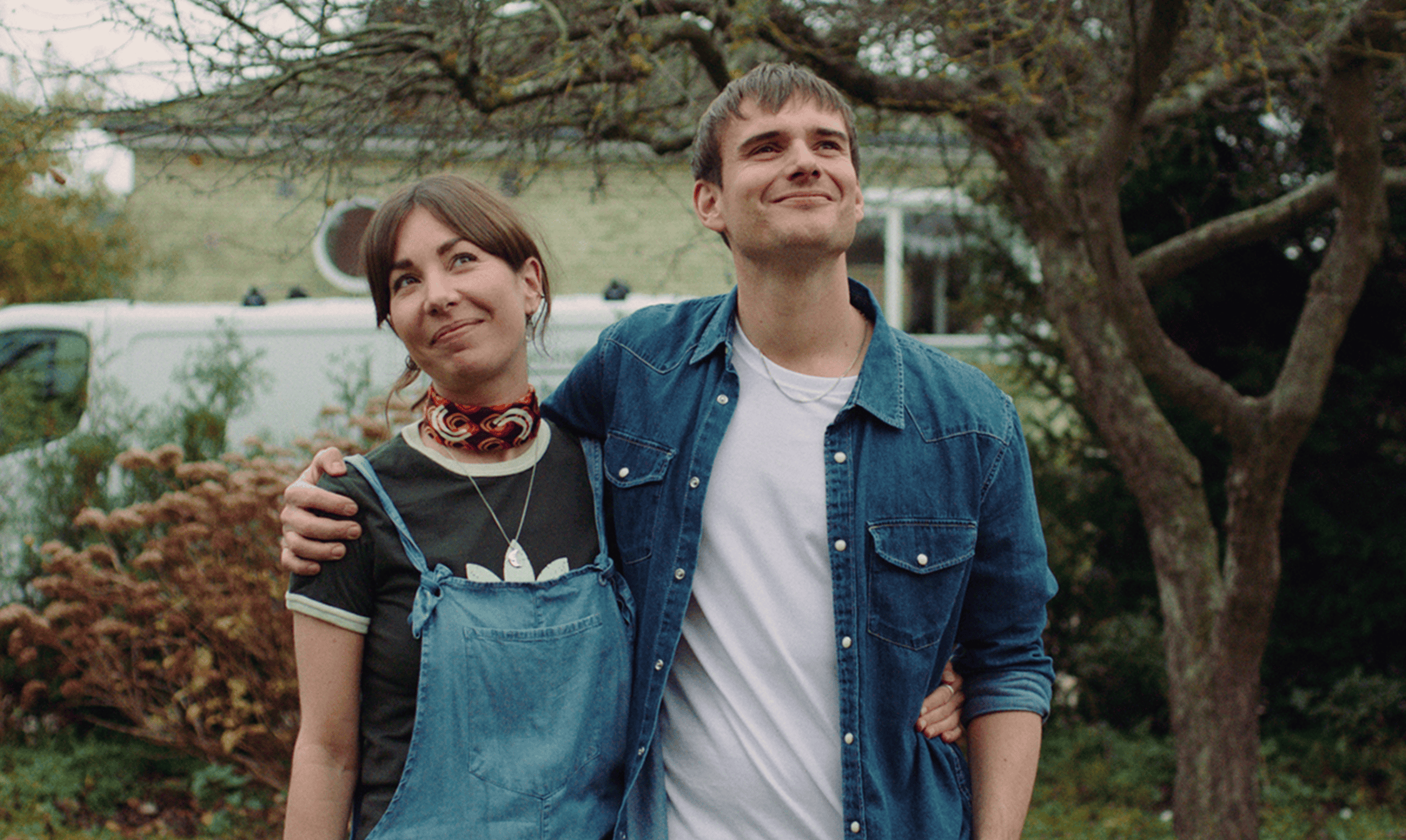 Vibrant outdoor shot of a smiling young couple embracing under a large tree, showcasing casual fashion and natural setting for professional film production.