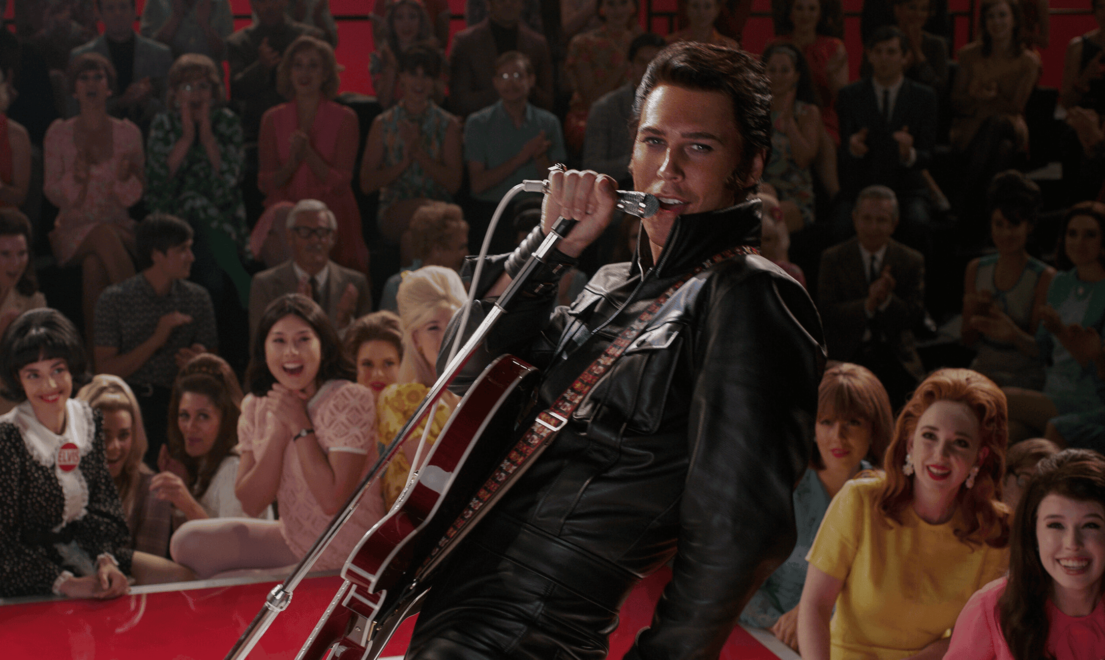 Brightly lit scene featuring a male performer with a guitar singing into a microphone on stage, surrounded by an animated audience of excited and happy women at a vintage-style event.