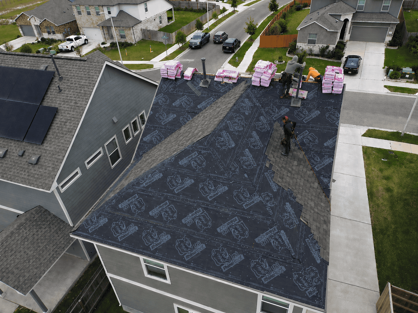 Residential house roof under construction with black RhinoRoof underlayment partially installed; workers placing asphalt shingles while bundles of shingles and tools are visible near the ridge, with neighboring homes in the background in San Antonio
