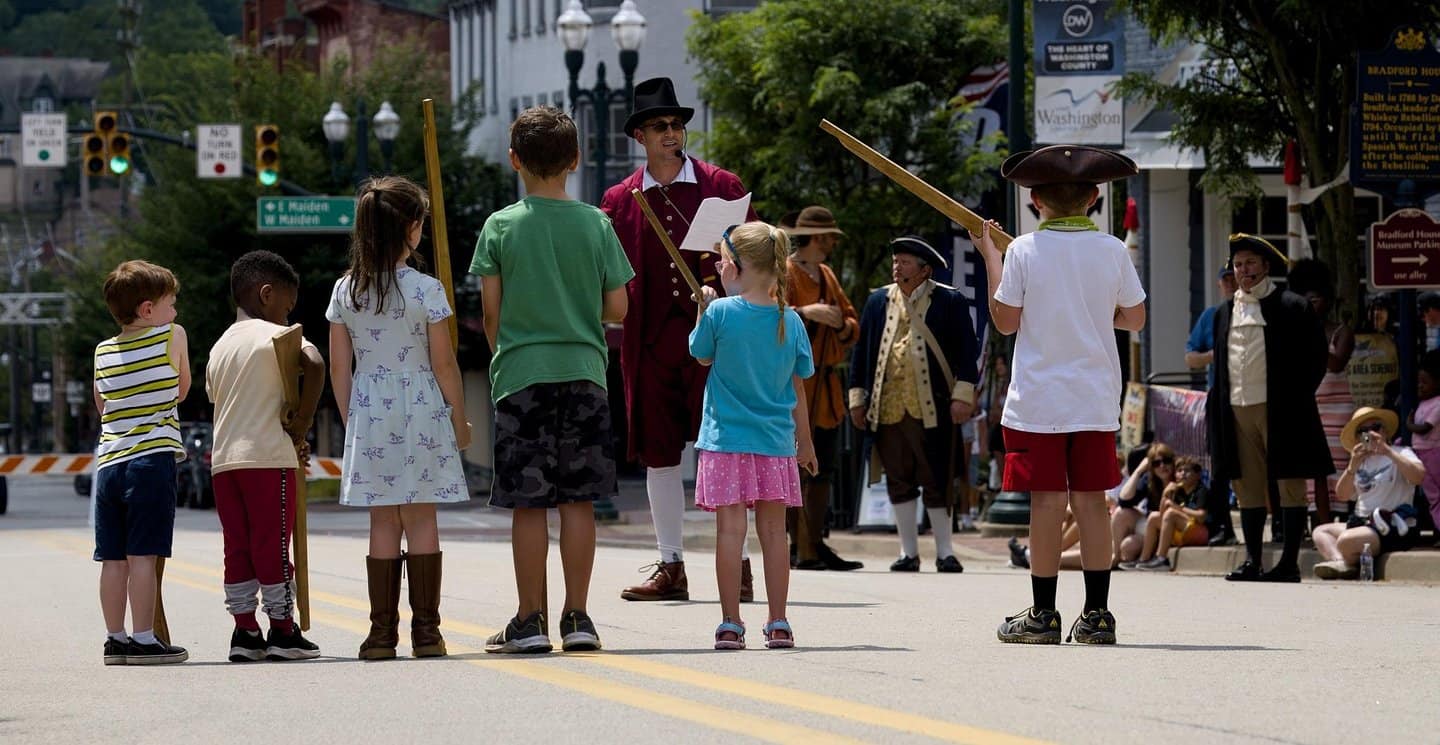Children and performers in historic costumes gather on a lively downtown street.