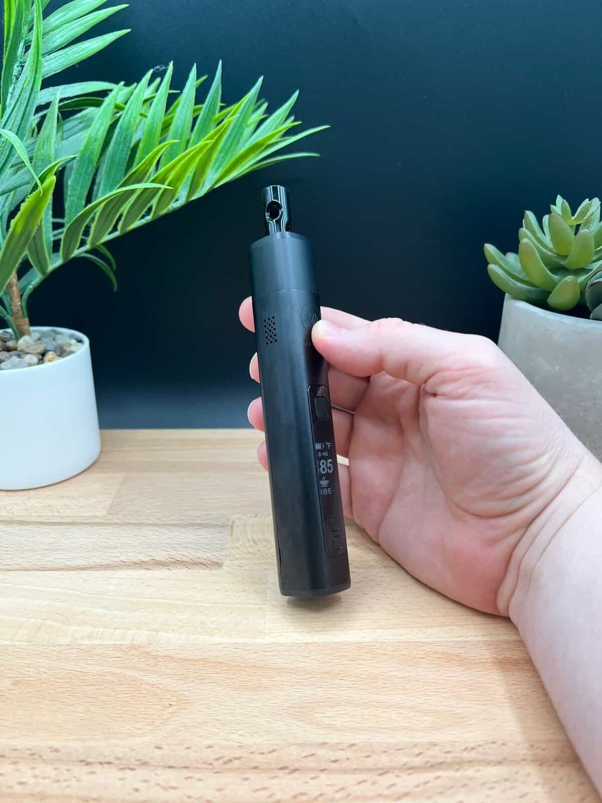Close-up of the XMAX V3 Pro dry herb vaporizer, showing visible fingerprints on its matte black aluminum body. The device is placed on a wooden surface with green plants in the background, highlighting its sleek design and real-world usage.