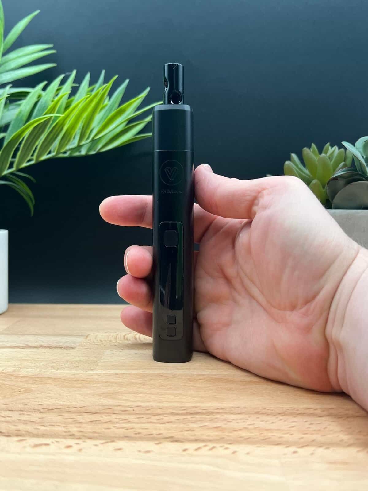 Close-up of the XMAX V3 Pro dry herb vaporizer held in hand, showing 385°F temperature on its digital OLED screen. The sleek black design contrasts with a wooden surface and potted plants in the background, emphasizing its modern aesthetics and functionality.