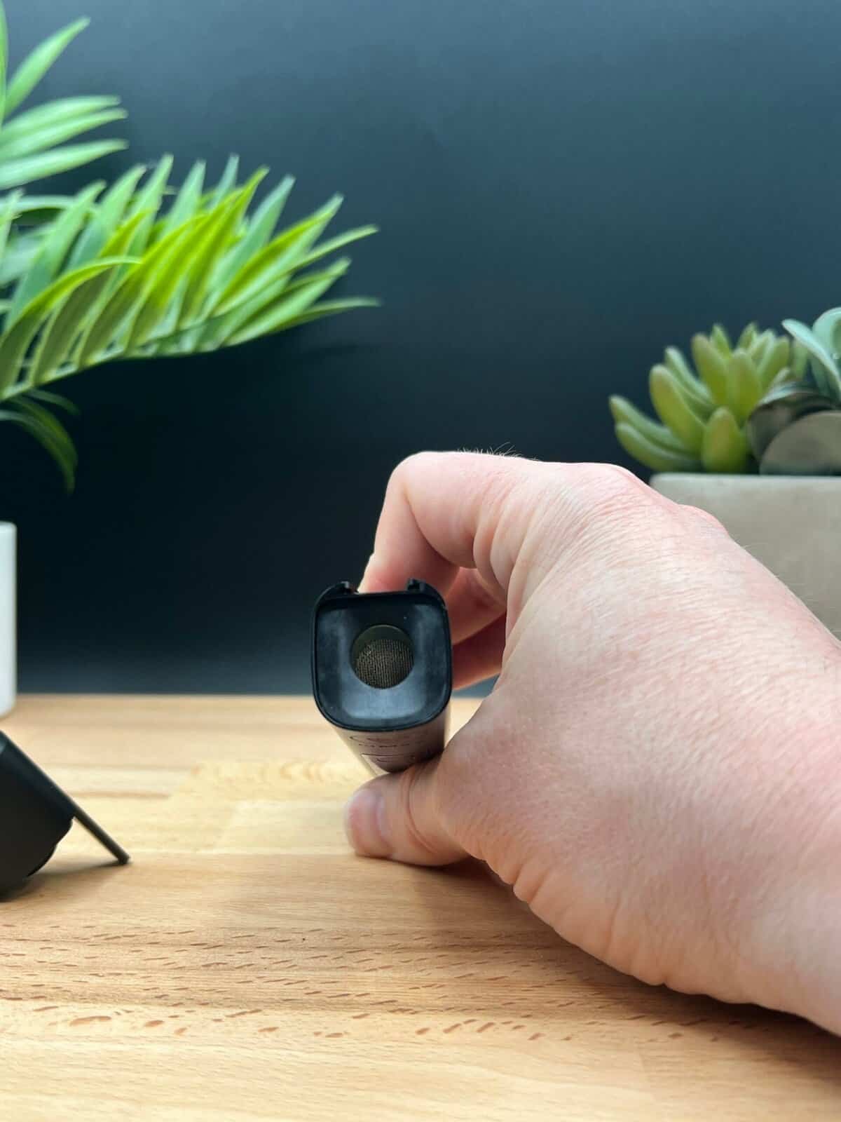 Close-up of the XMAX V3 Pro dry herb vaporizer with its stainless steel heating chamber fully exposed. The device is held vertically against a wooden surface with green plants in the background, emphasizing its sleek design and precision engineering.