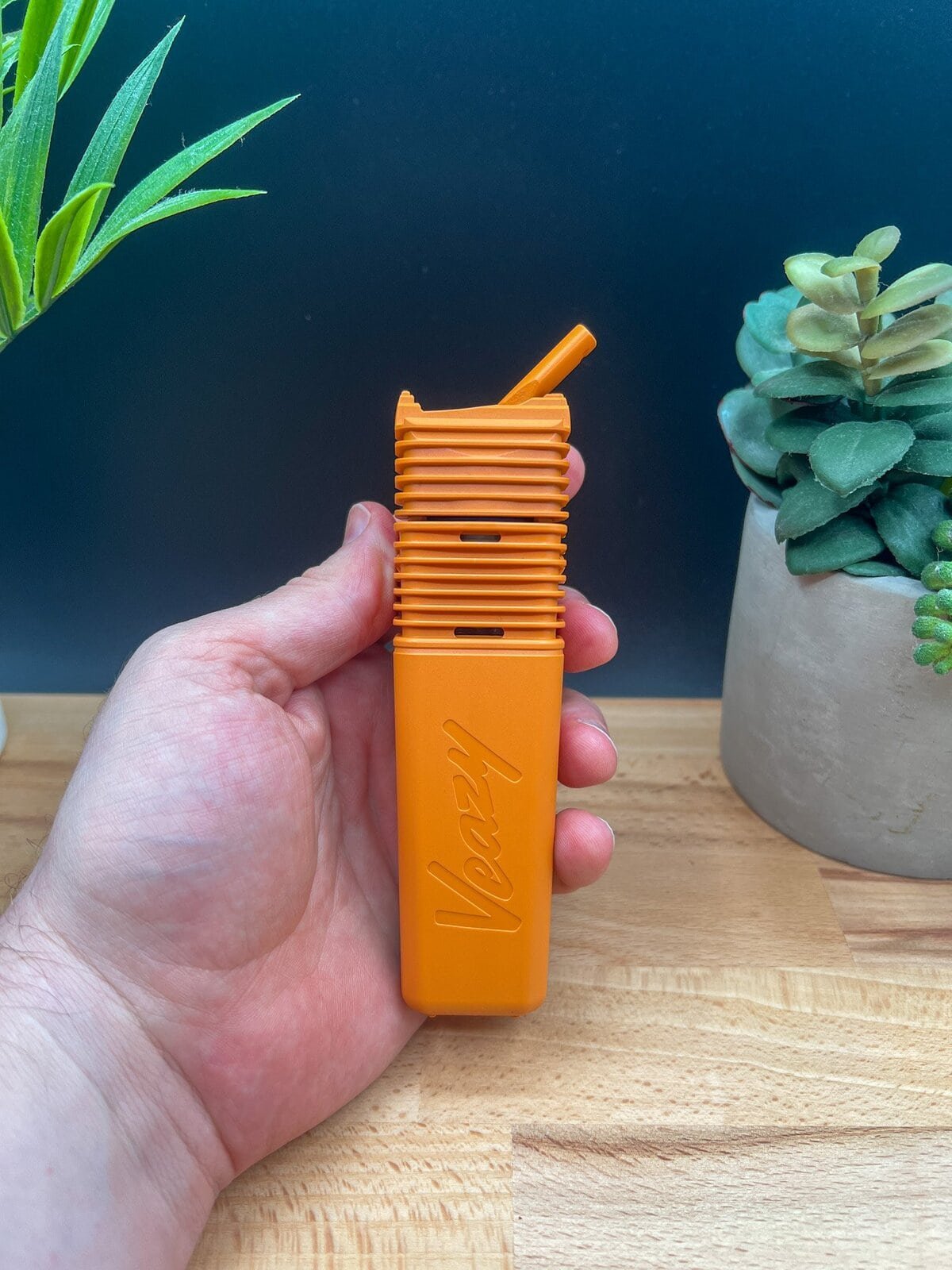 Hand holding the orange Storz & Bickel Veazy vaporizer with ribbed design and angled mouthpiece — featured in our Veazy Review.