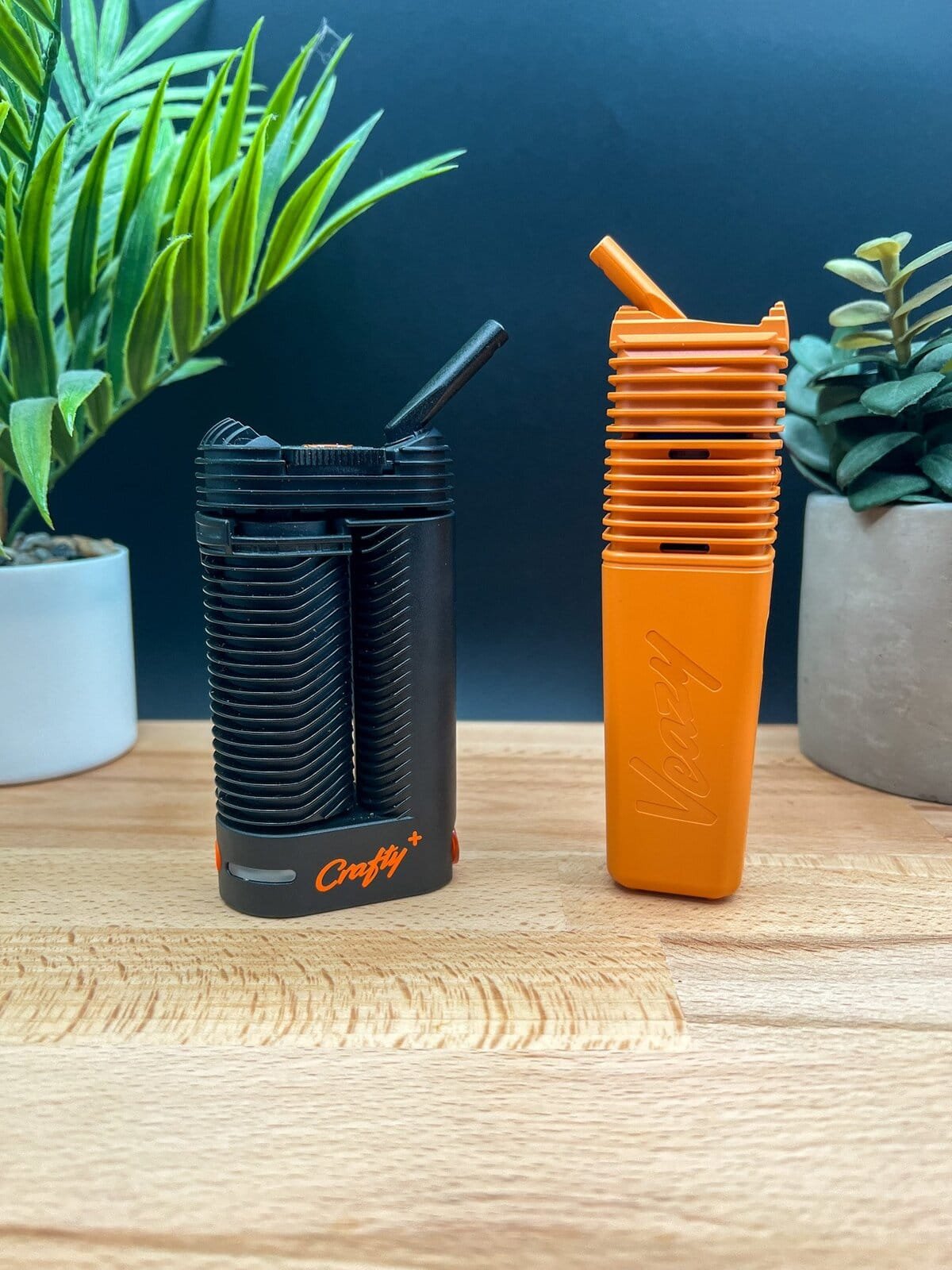 Side‑by‑side comparison of the black Crafty+ and orange Storz & Bickel Veazy vaporizers on a wooden surface — featured in our Veazy Review.