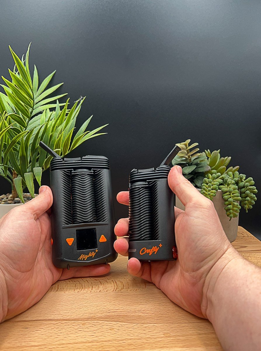 Hands holding the Mighty+ (Plus) and Arizer Solo 3 portable vaporizers side‑by‑side over a wooden table, showing design, size, and interface differences
