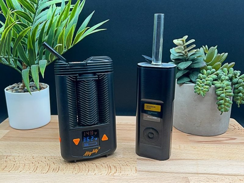 Side‑by‑side comparison of the Mighty+ (Plus) vs Arizer Solo 3 portable vaporizers on a wooden table, showing size, design, and display differences