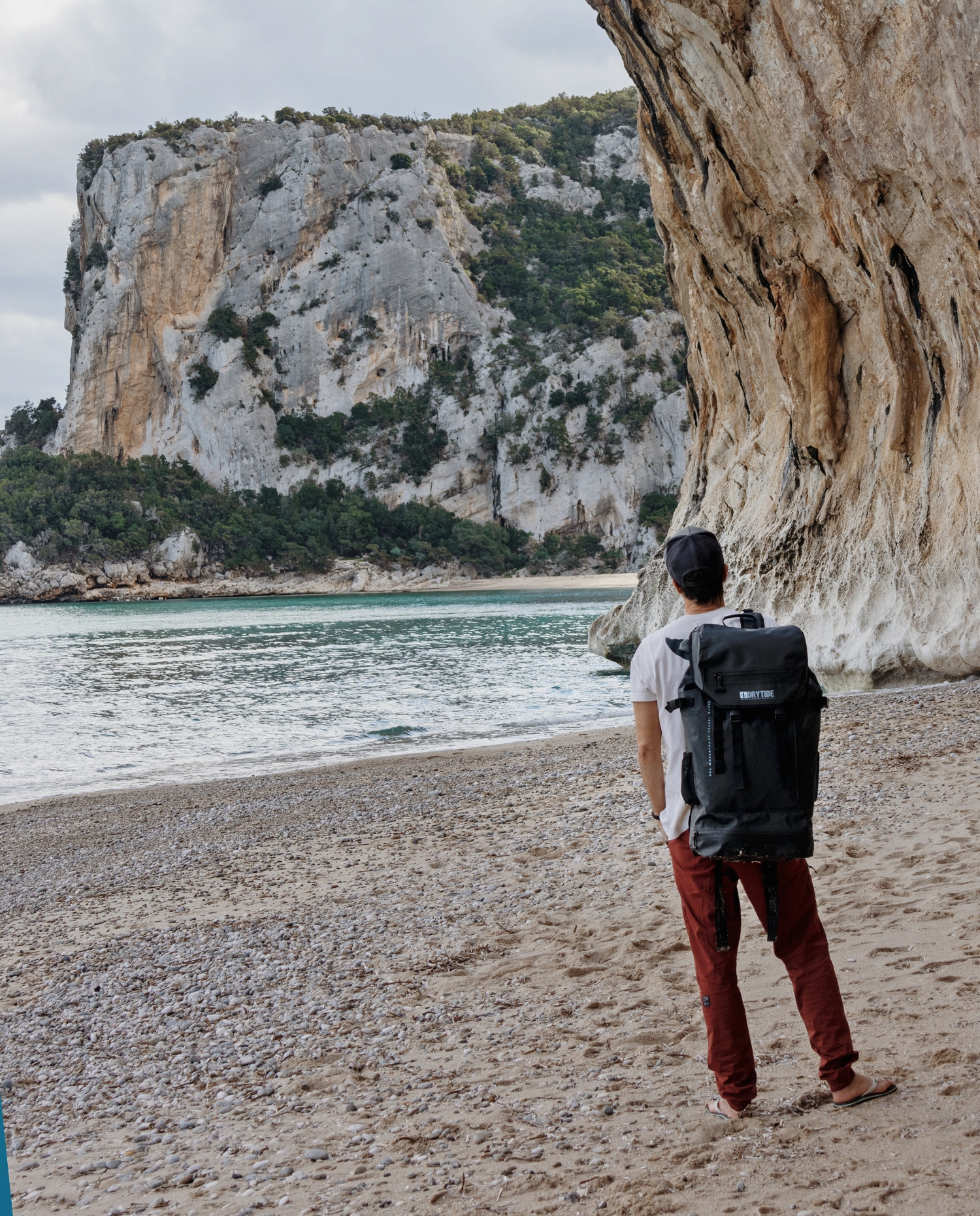 Person with DRYTIDE waterproof backpack on a beach with cliffs in the background.