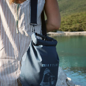 Waterproof dry bag with Drytide logo hanging from a person's shoulder near a river.