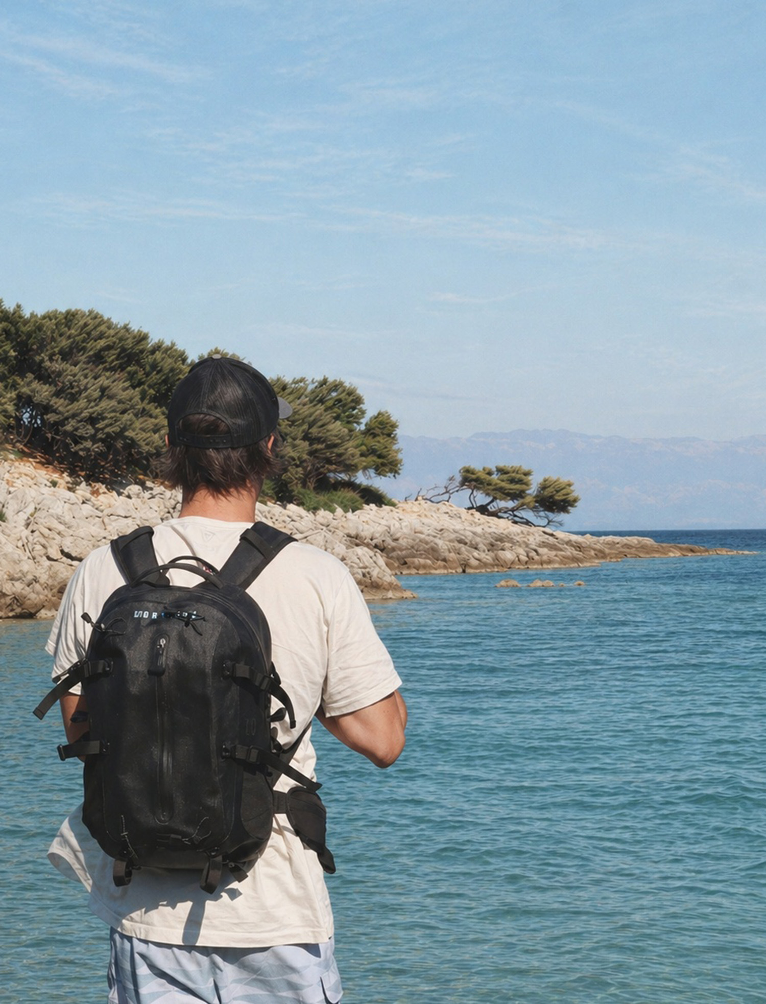 Waterproof backpack on hiker overlooking ocean shoreline with trees and blue sky.