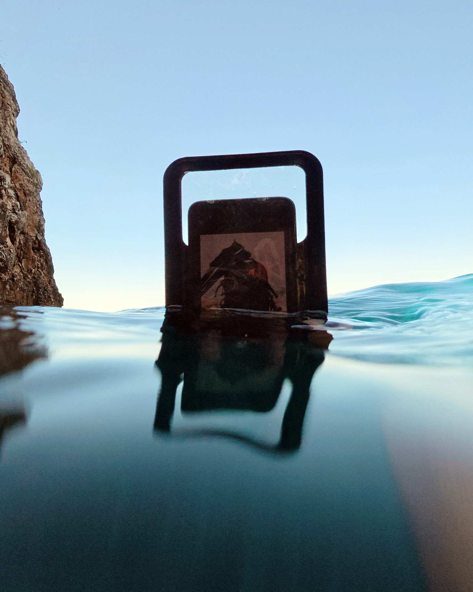 Waterproof phone pouch submerged in water near rocky shoreline.