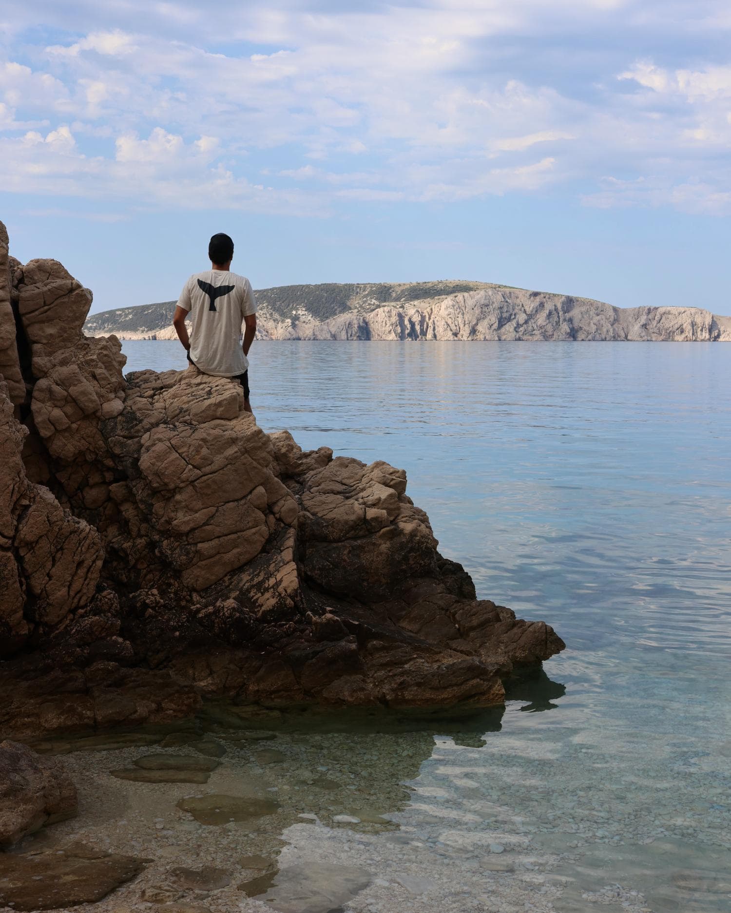 Man sitting on rocks by the water with a waterproof backpack, scenic coastal view.