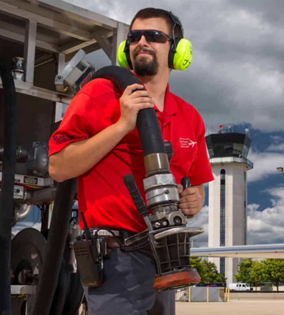 Man in red shirt, sunglasses, and yellow earmuffs holding a large fueling hose nozzle at an airport, with a control tower in the background.