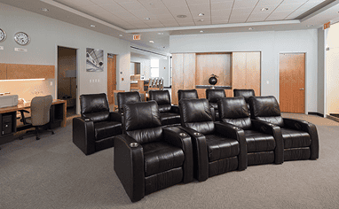An indoor lounge with two rows of dark brown leather recliners, a desk with a computer on the left, and wooden paneling in the background.