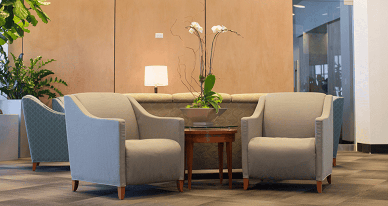 A modern lounge area with two grey armchairs, a wooden side table with orchids, and a sofa against a wood-paneled wall.